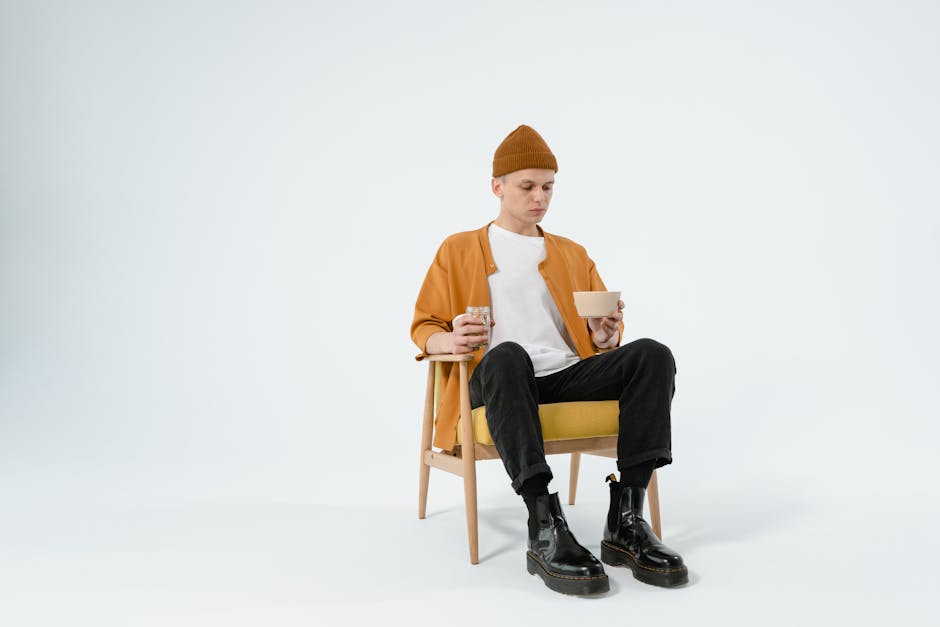 A man in trendy attire, holding a glass jar and bowl, sits in a stylish chair in a minimalist studio setup.