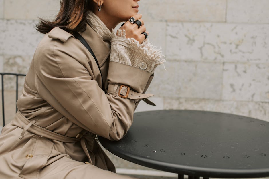 A fashionable woman in a beige trench coat sits outdoors at a table during autumn.